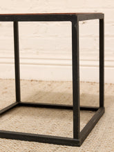 Load image into Gallery viewer, A close-up of a cube side table with a black metal frame and a dark brown wooden top, set on a beige textured rug against a white brick wall.
