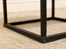 Load image into Gallery viewer, Close-up of a black metal table frame corner resting on a beige woven rug, with a light wall in the background.
