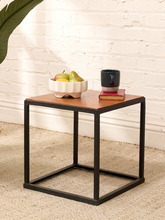 Load image into Gallery viewer, A square side table with a warm brown wood top and a black metal frame, styled with a bowl of fruit, a mug, and a book in front of a white brick wall.
