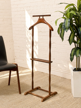 Load image into Gallery viewer, A wooden clothes rack with a hanger and shelf, standing in a room with a white brick wall, beige rug, a black chair on the left, and a green potted plant on the right.
