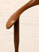 Load image into Gallery viewer, A close-up of a polished medium-brown wooden chair backrest showing the curved seat and vertical post against a light off-white brick wall background.
