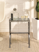 Load image into Gallery viewer, A small rectangular side table with a mirrored glass top and a silver metal frame, styled with a glass decanter, candle holder, and small cups in a bright room with a white brick wall and beige rug.
