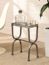 Load image into Gallery viewer, A small rectangular metal side table with a mirrored top and curved legs, styled in a room setting with a white brick wall, beige rug, a white chair, and a potted plant. The table is shown holding a glass decanter and two small candleholders.
