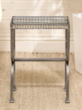 Load image into Gallery viewer, A rustic metal side table with a distressed silver finish, featuring a slatted top and curved legs, set on a woven jute rug against a white brick wall.
