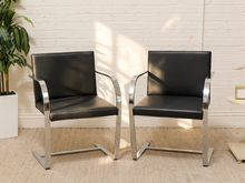 Load image into Gallery viewer, Two modern black leather armchairs with curved chrome metal frames, positioned side-by-side on a beige woven rug against a white brick wall.
