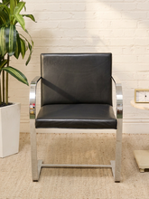 Load image into Gallery viewer, A modern black leather armchair with a polished chrome metal frame, set against a white brick wall background with a potted plant on the left.
