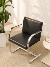 Load image into Gallery viewer, A modern black leather armchair with a chrome cantilever frame, positioned on a beige carpeted floor against a white brick wall, next to a potted plant.
