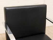 Load image into Gallery viewer, Close-up of a black upholstered chair back with a silver metal armrest, set against a white brick wall and beige carpet.
