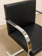 Load image into Gallery viewer, A close-up of a modern black leather or faux-leather chair with a curved polished chrome armrest and matching metal base, shown on a woven beige rug against a white wall.
