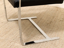 Load image into Gallery viewer, Close-up of a modern chair leg with a polished silver metal T-shaped base resting on a beige textured rug.
