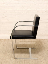 Load image into Gallery viewer, A modern cantilever chair with a black leather seat and backrest, supported by a polished chrome steel frame, set against a white brick wall and a beige woven rug.
