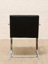 Load image into Gallery viewer, A modern black leather chair with a chrome cantilever steel base, shown from the back against a white brick wall and beige rug.
