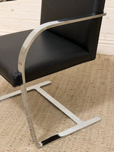 Load image into Gallery viewer, A modern black leather chair with a curved chrome metal cantilever base, shown on a woven jute rug against a white brick wall.
