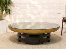 Load image into Gallery viewer, A round coffee table with a thick glass top, a light natural wood rim, and a black geometric pedestal base, set on a beige rug against a white brick wall.

