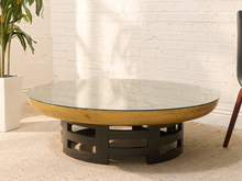 Load image into Gallery viewer, A round coffee table with a clear glass top, a light natural wood rim, and a black metal geometric base, set in a room with a white brick wall and beige carpet.
