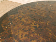 Load image into Gallery viewer, A close-up of a round table with a distressed, mottled brown and black wood-grain top, showing peeling paint or wear on the surface.

