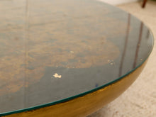 Load image into Gallery viewer, Close-up of a round coffee table with a distressed gold metal base and a clear glass top showing a mottled brown surface underneath.
