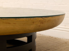 Load image into Gallery viewer, Close-up of a round table with a distressed gold-painted wooden rim, a clear glass top, and a dark metal base against a white brick wall and beige rug.
