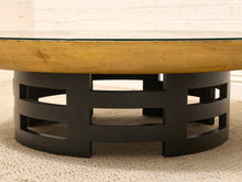 Load image into Gallery viewer, A round coffee table with a light natural wood top, a clear glass upper rim, and a black metal geometric base, shown on a textured beige rug against a white brick wall.
