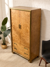 Load image into Gallery viewer, A tall mid-century modern armoire cabinet with a warm golden-brown burl wood finish and brass trim, shown in a styled room setting next to a potted plant and a black chair.
