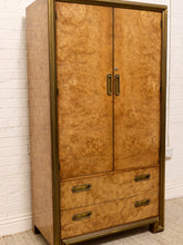 Load image into Gallery viewer, A tall two-door wardrobe or armoire with a prominent burl wood grain finish in warm golden-brown tones, framed in brass/gold-toned metal, featuring two upper doors with vertical handles and two lower drawers with horizontal handles, set against a white brick wall.
