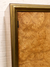 Load image into Gallery viewer, Close-up of a cabinet door featuring dramatic swirling burl wood grain in warm golden-brown tones, framed by a wide ornate gold-toned border against a white brick wall.
