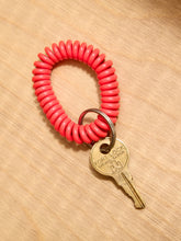 Load image into Gallery viewer, A bright coral pink coiled spring keychain with a metal key attached, displayed on a light wood grain surface.
