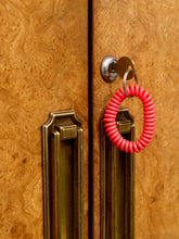 Load image into Gallery viewer, A bright red coiled door security keychain hangs from a brass keyhole on a wood-paneled door with brass handles.
