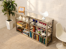 Load image into Gallery viewer, A three-tier mirrored bookcase with a glass top, styled with books, glasses, and decorative items in a bright room with a white brick wall, a potted plant, and a white chair.
