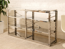 Load image into Gallery viewer, A modern three-compartment shelving unit with a polished chrome metal frame and clear glass shelves, set against a white brick wall and beige carpet.

