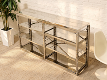 Load image into Gallery viewer, A modern three-tier metal shelving unit with a polished chrome frame and clear glass shelves, shown in a styled room setting with a white brick wall, beige carpet, a potted plant, and a white chair.
