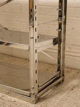 Load image into Gallery viewer, A close-up of a polished chrome metal shelving unit with three mirrored shelves, set against a white brick wall and beige carpet.
