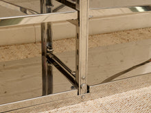 Load image into Gallery viewer, Close-up of a polished stainless steel shelving frame with mirrored panels and visible screws and support brackets, set against a beige brick wall and carpeted floor.
