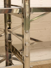 Load image into Gallery viewer, Close-up of a polished stainless steel shelving unit with adjustable pegboard-style side rails and visible mounting bolts, set against a light brick wall background.
