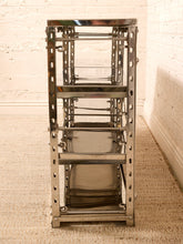 Load image into Gallery viewer, A tall, narrow stainless steel shelving unit with multiple tiers and side peg holes, standing on a textured beige rug against a white brick wall.
