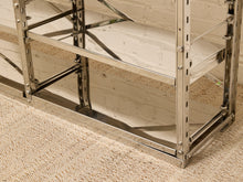 Load image into Gallery viewer, A close-up of a polished stainless steel industrial shelving unit with three tiers, set against a white brick wall and a beige textured carpet.
