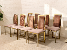 Load image into Gallery viewer, A set of eight dining chairs with blush pink velvet upholstered seats and backs, framed in polished gold metal, arranged against a white brick wall.

