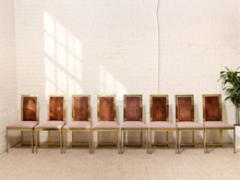 Load image into Gallery viewer, A row of eight matching dining chairs with blush pink upholstered seats and backs, bronze rectangular panels, and gold metal frames, arranged against a white brick wall.
