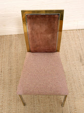 Load image into Gallery viewer, A mid-century modern dining chair featuring a textured mauve-pink upholstered seat and backrest, framed by a polished gold-tone metal border and legs, set on a woven jute rug.
