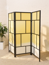 Load image into Gallery viewer, A three-panel folding room divider with a black metal frame and translucent yellow and white panels, styled in a bright room with a white brick wall, beige carpet, and a potted plant.
