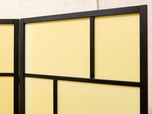 Load image into Gallery viewer, A close-up of a folding screen or room divider with pale yellow panels and a black metal frame, shown against a white brick wall.
