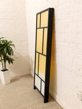 Load image into Gallery viewer, A tall, narrow black-framed mirror with a warm yellow-to-white gradient reflection, leaning against a white brick wall on a beige carpet next to a potted plant.
