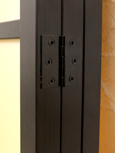 Load image into Gallery viewer, Close-up of a pair of black double-core hinges installed on dark wood doors, with a cream wall on the left and a textured orange wall on the right.
