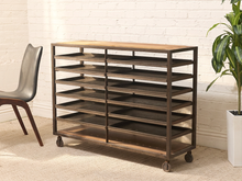 Load image into Gallery viewer, A 7-tier rolling shoe rack with a dark metal frame, light wood top and shelves, and caster wheels, shown in a styled room setting with a white brick wall, a chair, and a potted plant.
