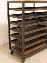Load image into Gallery viewer, A dark brown rolling bakery or display rack with multiple tiers and caster wheels, shown in a room setting against a white brick wall and textured rug.
