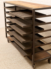 Load image into Gallery viewer, A rustic industrial rolling storage rack with a light wood top, dark metal frame, and multiple dark wood shelves on casters, shown in an indoor setting on a textured rug.

