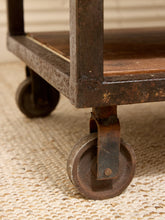 Load image into Gallery viewer, Close-up of the lower corner of a rustic metal rolling cart with a wooden shelf and casters, resting on a woven beige rug.
