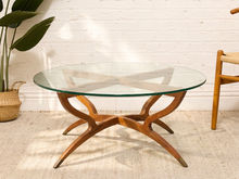 Load image into Gallery viewer, Vintage mid-century modern round glass coffee table with sculptural curved wood base, crossed supports, and brass-tone feet.
