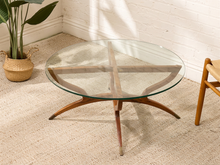 Load image into Gallery viewer, A mid-century modern round coffee table with a clear glass top and a wooden starburst base, set on a beige textured rug in a room with a white brick wall and a potted plant.

