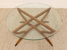 Load image into Gallery viewer, A round glass-top coffee table with a mid-century wooden starburst base and angled legs, shown on a woven rug.
